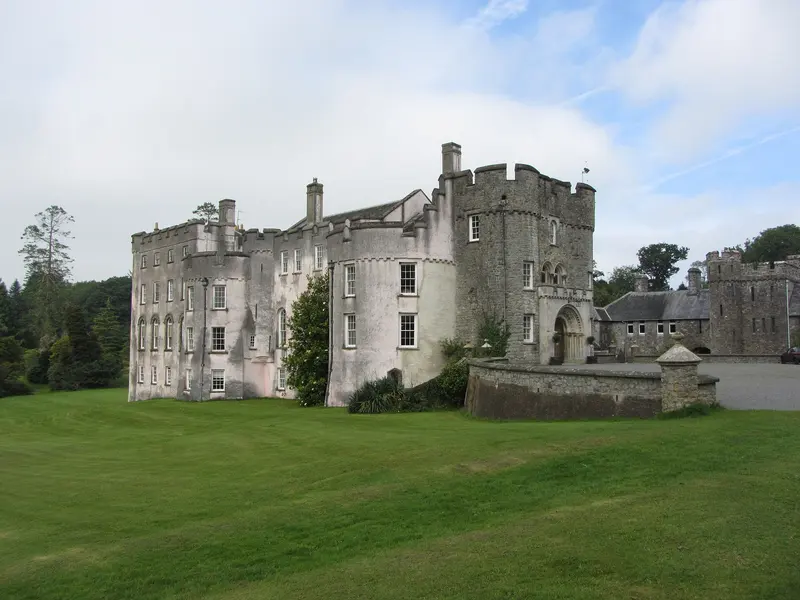 The pale battlemented walls and round towers of Picton Castle beside a sweeping lawn, with outbuildings and trees under a cloudy sky