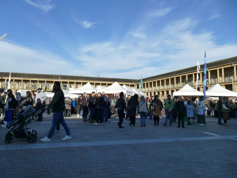 Crowds of shoppers among white market gazebos in the sunlit stone courtyard of the Piece Hall, ringed by its two-storey colonnades under a blue sky