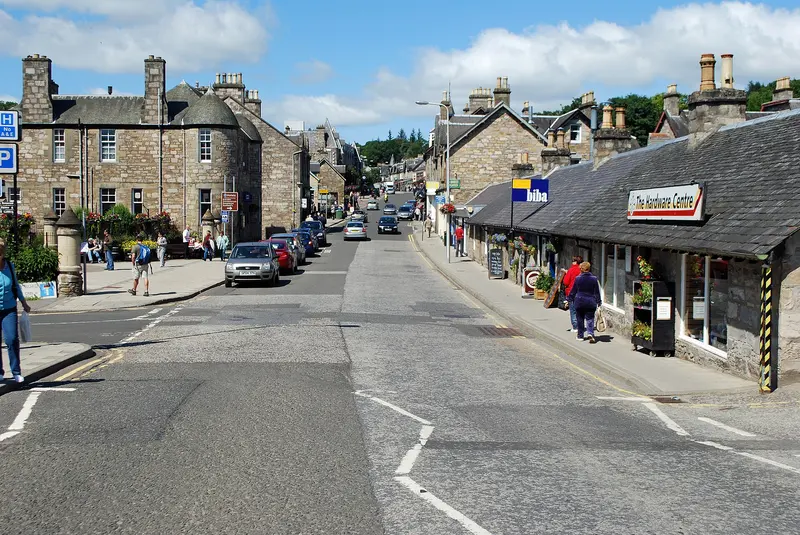 Pitlochry's Atholl Road on a sunny day, lined with grey stone Victorian buildings and low shops, with wooded hills beyond