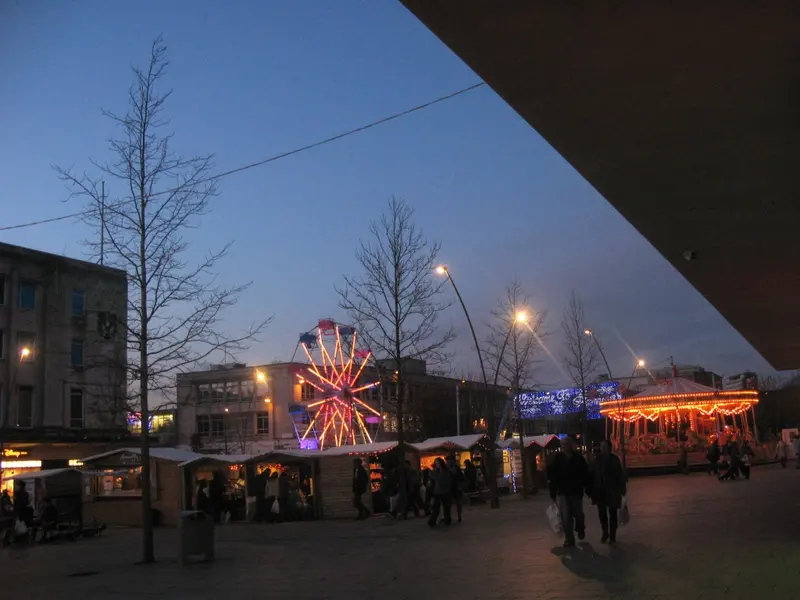 Wooden Christmas market chalets, a lit carousel and a big wheel on a Plymouth city-centre street at dusk, with shoppers passing