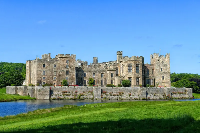 The towers and battlemented walls of Raby Castle above a moat-like pond and green lawns under a clear blue sky