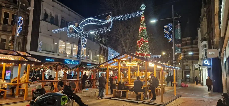 Wooden market huts and covered seating strung with warm lights on Broad Street at night, beneath a tall Christmas-tree light sculpture and festive street decorations