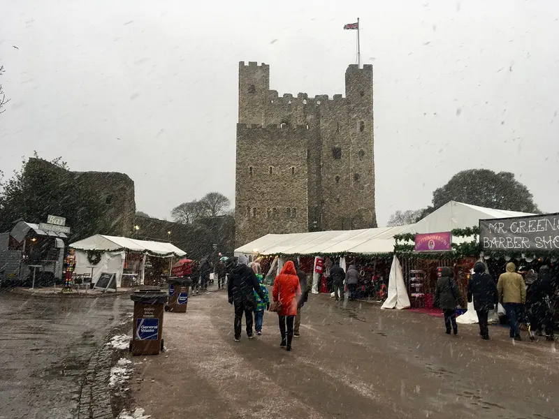 Snow falling on white market marquees and stalls in Rochester Castle Gardens, with shoppers in coats and the great stone keep of Rochester Castle behind