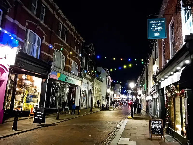 Rochester's historic High Street at night, with lit shop windows, a garlanded doorway and strings of coloured lights across the narrow street
