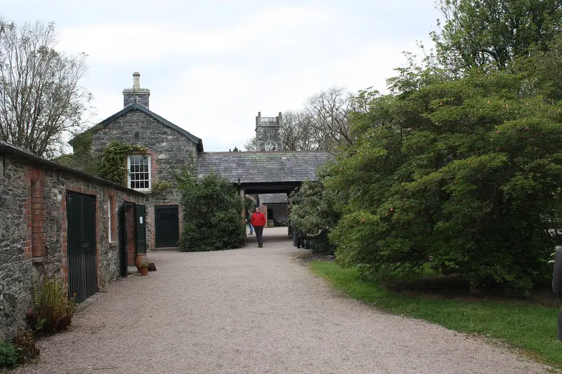 Old stone outbuildings with green doors along a gravel yard at Rowallane Garden, with a small clock tower beyond and leafy shrubs