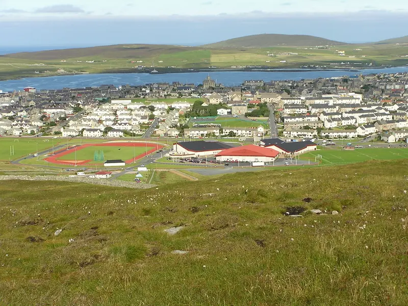 A hillside view over Lerwick, with the red-roofed Clickimin Leisure Complex and its running track among houses, and the sea and low hills beyond
