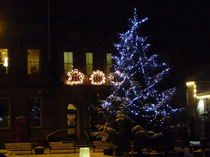 A tall Christmas tree strung with blue-white lights in front of Skipton Town Hall at night, with light decorations on the building and a dusting of snow