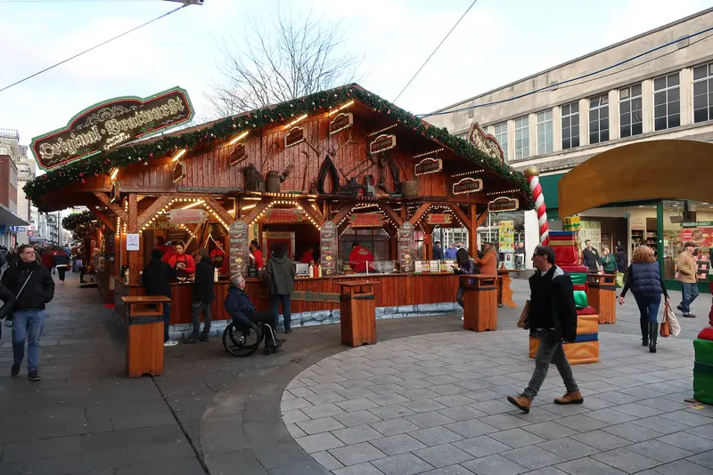 A large carved wooden bratwurst stall decorated with garlands on Above Bar Street, with shoppers walking past in winter sunshine