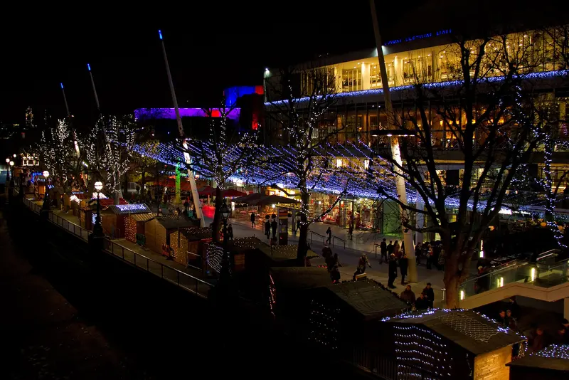 Wooden market chalets edged with white fairy lights along the riverside walk at night, beneath trees strung with lights and the glowing Royal Festival Hall