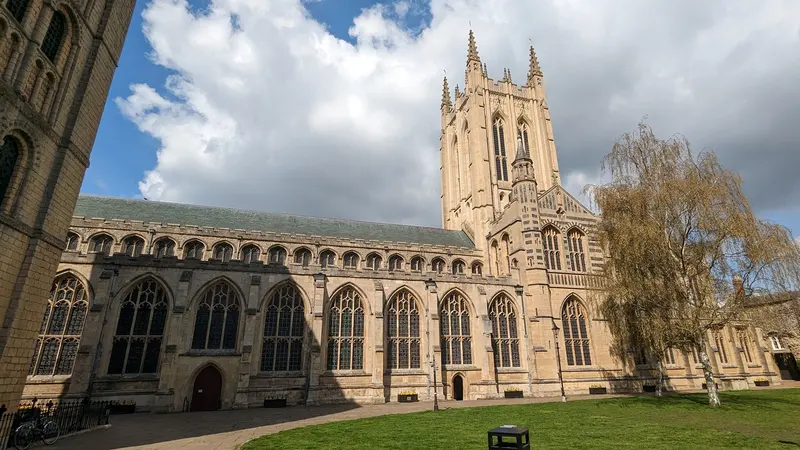 The pale stone St Edmundsbury Cathedral with its tall pinnacled tower and long nave, beside a lawn and a willow tree under a blue sky with clouds