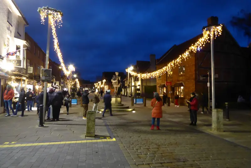 Henley Street in Stratford-upon-Avon at dusk, with garlands of warm lights strung from lamp posts along timber-framed buildings and people strolling on the paving