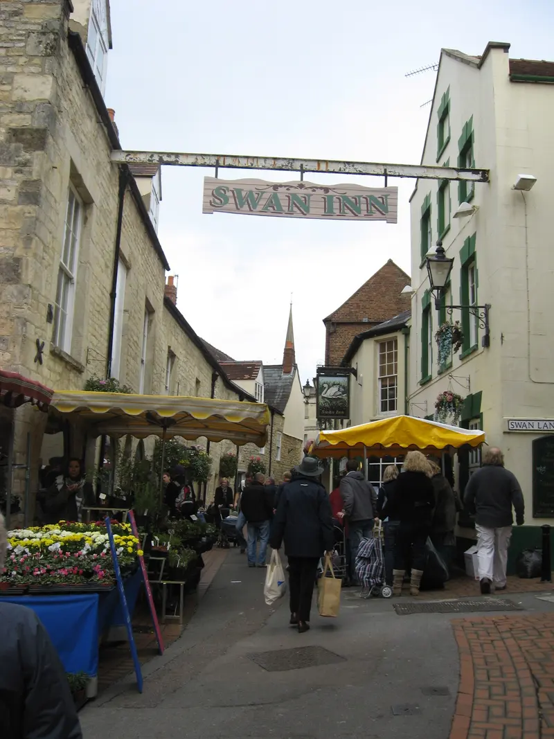 Market stalls with yellow awnings and buckets of flowers in a narrow stone-built lane in Stroud, under the hanging sign of the Swan Inn
