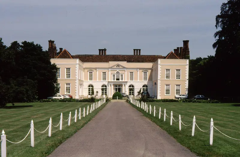 The pale classical front of Hintlesham Hall at the end of a long driveway lined with white posts and chains, between mown lawns and trees