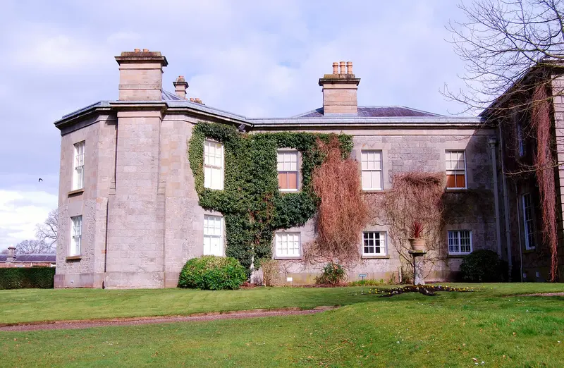 The pale stone neoclassical house of The Argory, partly covered in ivy and red creeper, beside a lawn under a soft blue sky
