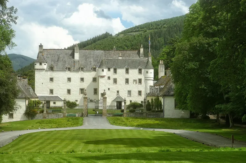 The white-harled Traquair House with its steep slate roofs and small turrets, behind gate piers and a wide striped lawn, with wooded hills behind