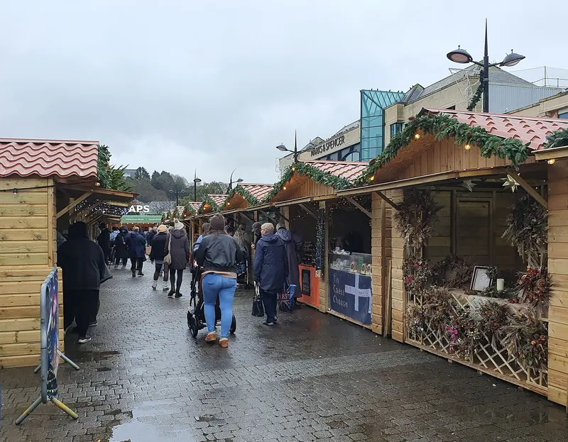 Wooden chalets with red-tiled roofs, garlands and wreaths line Lemon Quay in Truro, with shoppers in winter coats walking between them on a grey day