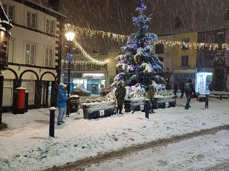 Snow falling at night in Ulverston town centre, with a lit Christmas tree, strings of white lights and old shopfronts