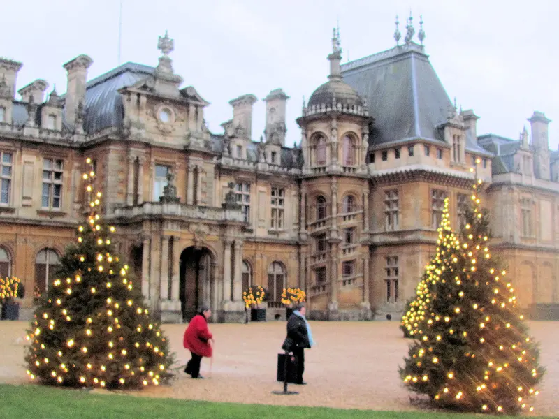 Two Christmas trees covered in warm lights in front of the ornate French-château-style facade of Waddesdon Manor, with visitors crossing the forecourt