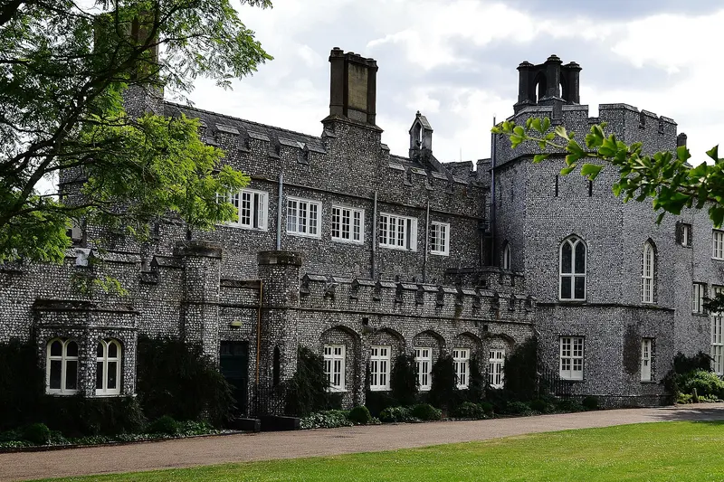 The flint-faced, battlemented front of West Dean House, now West Dean College, with tall chimneys and Gothic windows beside a lawn