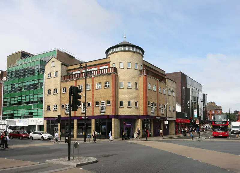 A busy junction in Wimbledon town centre, with a turreted brick-and-stone corner building, modern glass offices and a red London bus