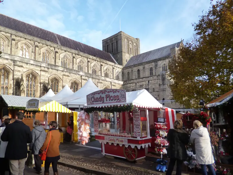 A red-and-white candy floss cart and wooden market chalets in the Cathedral Close, with the long nave and tower of Winchester Cathedral behind under a blue sky