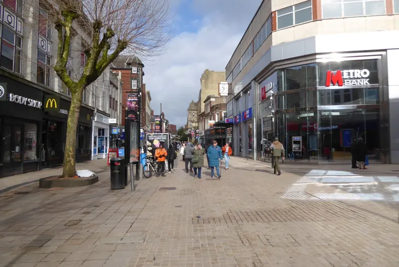 The pedestrianised Dudley Street in Wolverhampton city centre, lined with shops and a pollarded tree, with shoppers walking towards the old buildings at the far end
