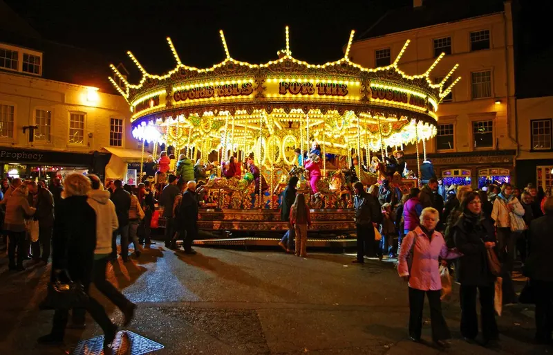A brightly lit gallopers carousel at night in Cornmarket, Worcester, surrounded by crowds of visitors in winter coats