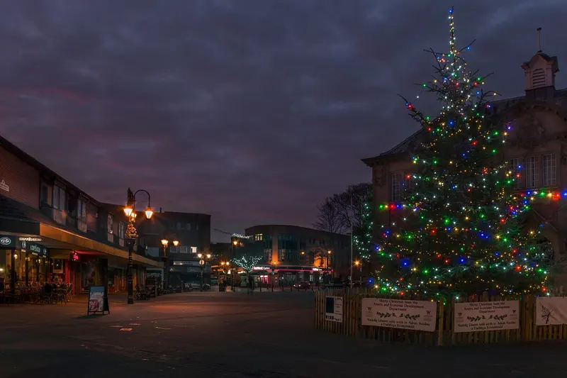 A tall Christmas tree strung with coloured lights in Queen's Square, Wrexham, at dusk, with lamp-lit shopfronts and a cloudy purple sky