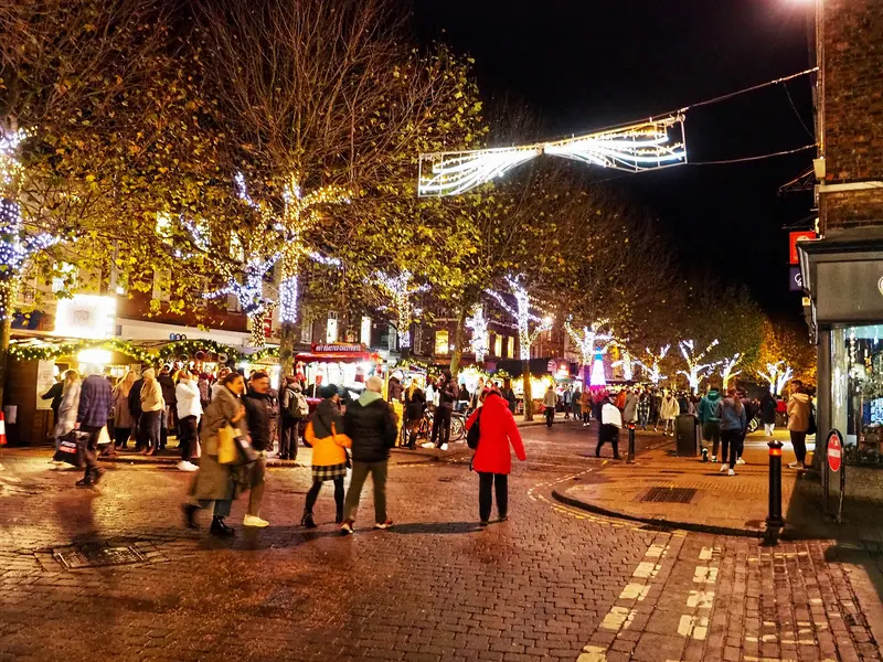 Parliament Street in York at night, with market chalets under trees wrapped in white lights, a light decoration strung across the street and shoppers on the cobbles