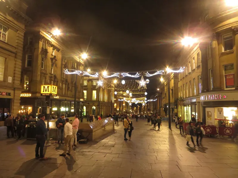 Strings of Christmas lights and stars across the top of Grey Street in Newcastle at night, with grand stone buildings and people out in the evening
