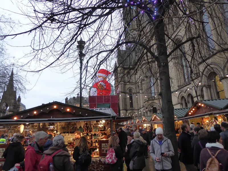Crowds among lit wooden Christmas market chalets, with a giant Santa figure, bare trees and the Gothic Manchester Town Hall behind
