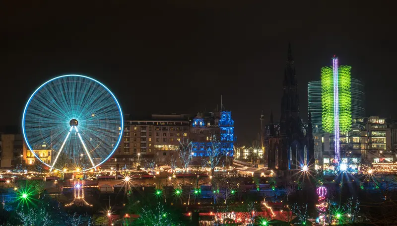 Edinburgh's Christmas fair at night, with a big wheel lit in blue, rows of glowing market stalls and the dark spire of the Scott Monument