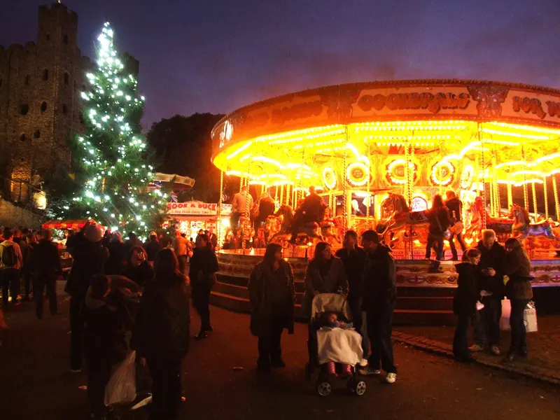 A glowing gallopers carousel and a Christmas tree covered in white lights at dusk, with visitors gathered below the walls of Rochester Castle