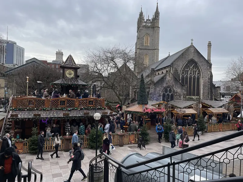 A two-storey wooden Christmas bar with a clock tower and festive chalets on Working Street, Cardiff, with St John's Church tower behind