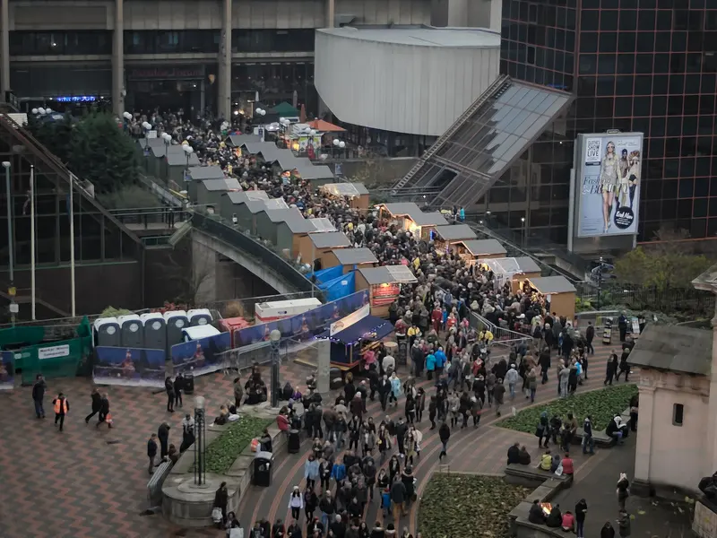 Crowds walking between rows of Christmas market chalets that line the paved squares of Birmingham city centre, seen from above