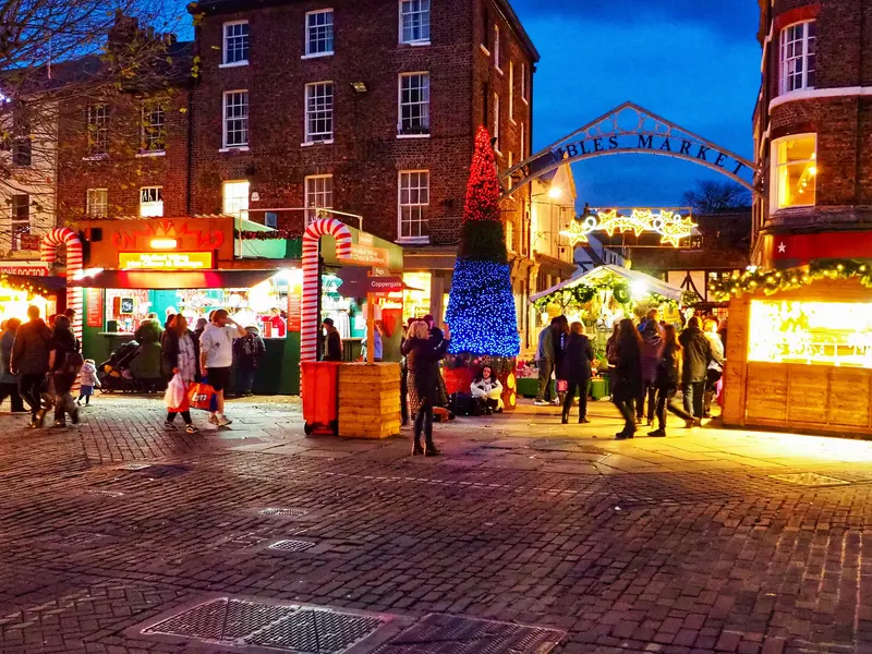 Brightly lit market stalls, candy-cane arches and a tall blue-and-red light-up tree in a cobbled York square at dusk, under the Shambles Market sign