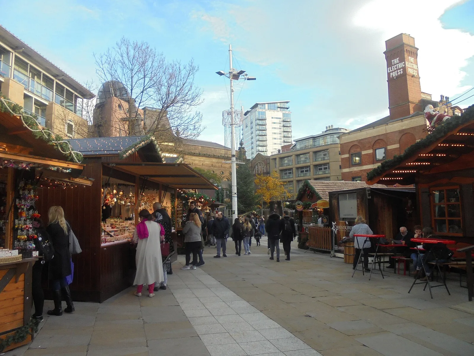 Wooden Christmas market chalets decorated with garlands and lights on a sunny paved square, with shoppers browsing stalls and a Santa sleigh on a roof under a blue sky